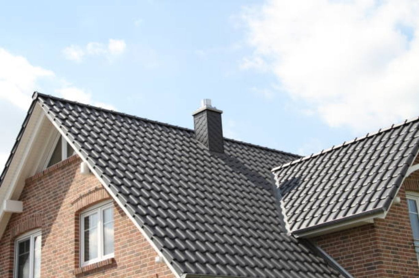 Dark glazed tile roof with a dark chimney and brick house walls under a partly cloudy sky