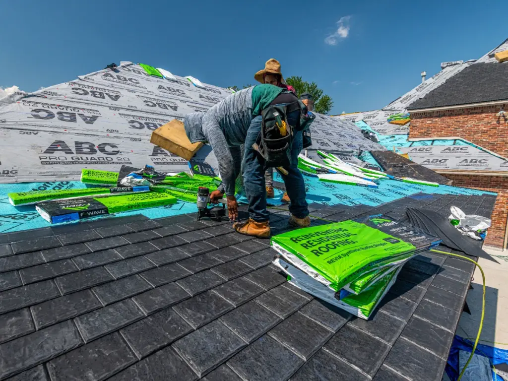 Roofer kneeling on a partly shingled roof, with rolls of underlayment and bundles of shingles beside him