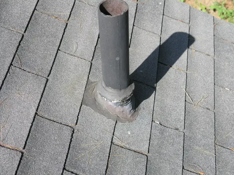 Worn, patched flashing at the base of a plumbing vent pipe on a gray shingle roof